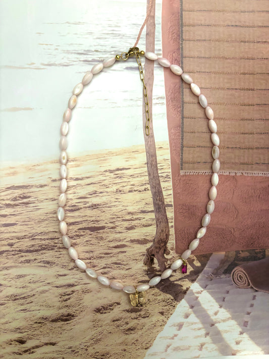 Blush pink coral bead necklace with gold butterfly charm and hot pink crystal accent, styled on a sandy beach backdrop with ocean in the distance. Features a 18k gold-plated clasp and extender chain. Handmade by Soul Wave Beach Co, this soft and coastal necklace offers a feminine, beachy touch—perfect for summer layering or dreamy resort styling.